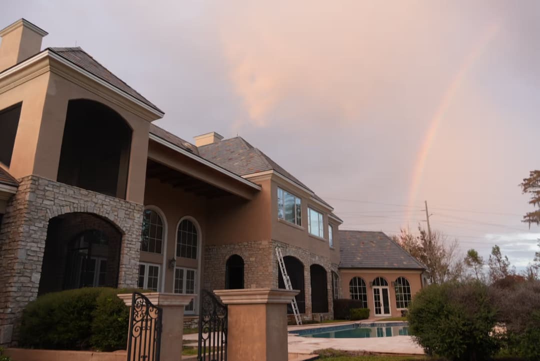Luxury home with stone accents, pool, and rainbow in the sky at sunset.
