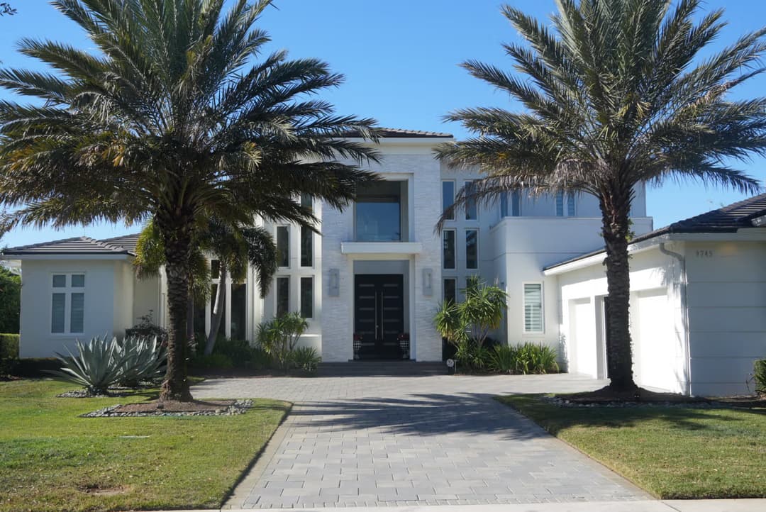 Modern luxury home with palm trees, stone pathway, and white facade against a blue sky.