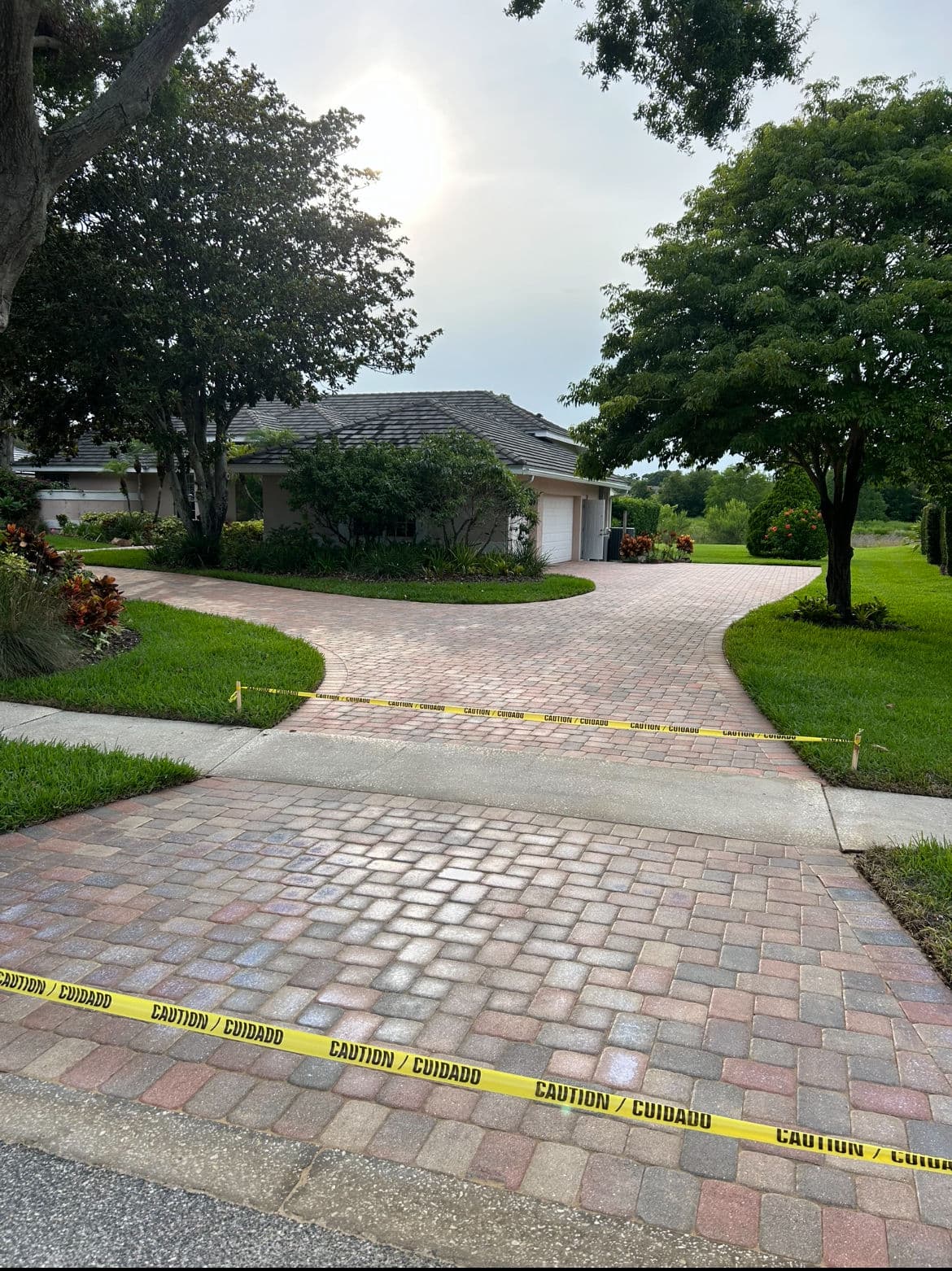 Caution tape blocking brick driveway near house with trees and cloudy sky.